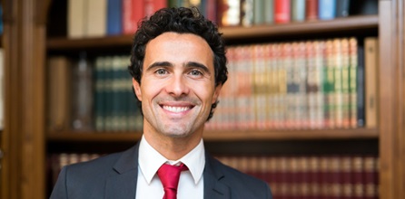 A smiling man in a suit in front of bookshelves.