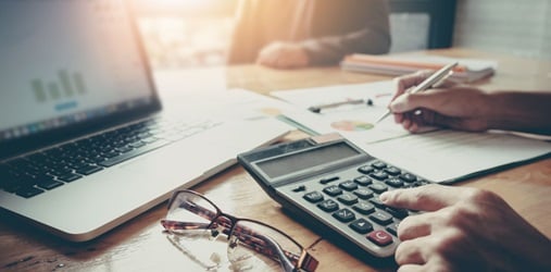 A closeup of a calculator, a computer, and a person writing their tax deductions on a paper.